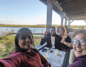 Four people sit at a table on an outside patio at dinner. There is greenery and water in the background. All four people are smiling at the camera.